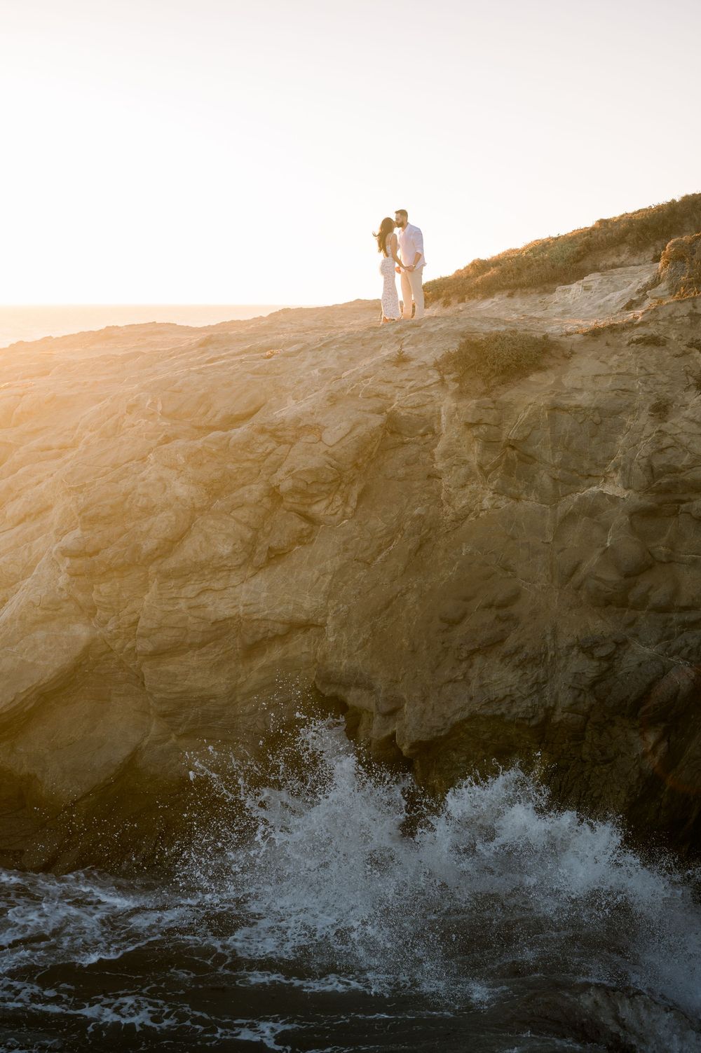 Ocean waves crash against rocky cliffs at sunset while silhouettes stand atop the coastal overlook.