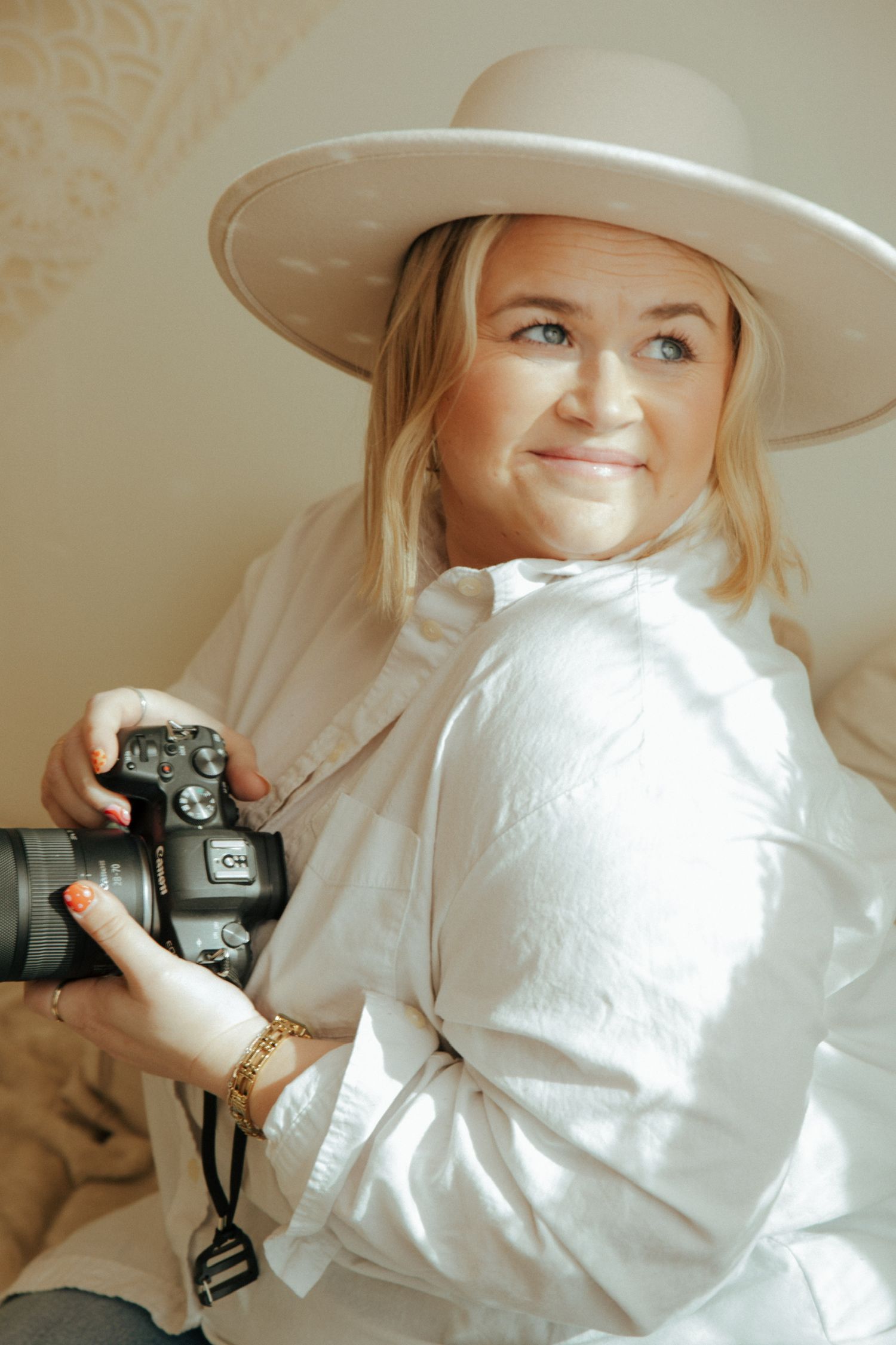 Person wearing a white blouse and beige hat poses playfully with a vintage camera in a light-filled room.