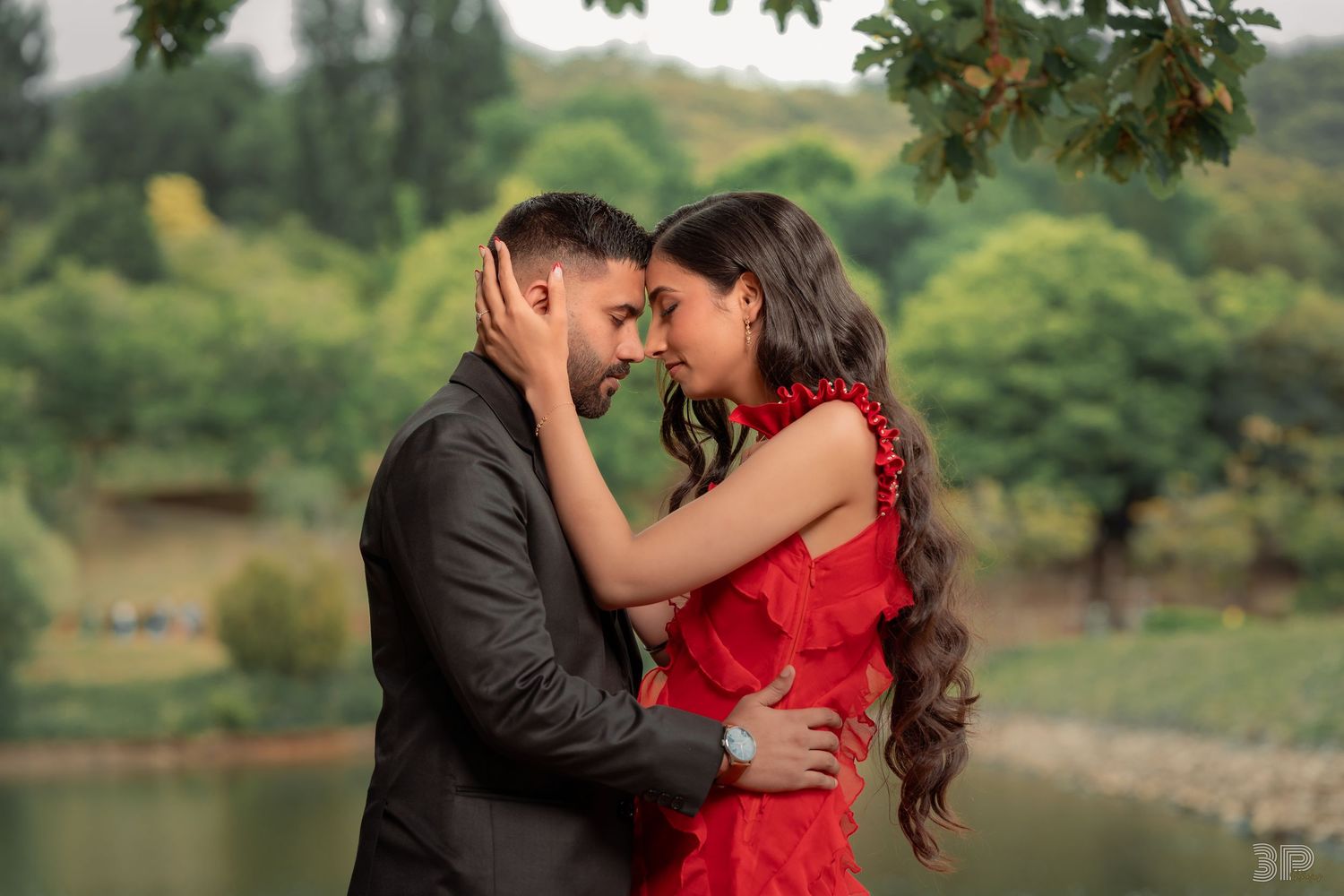 A couple in formal attire shares an intimate moment by a scenic lake with trees in the background.