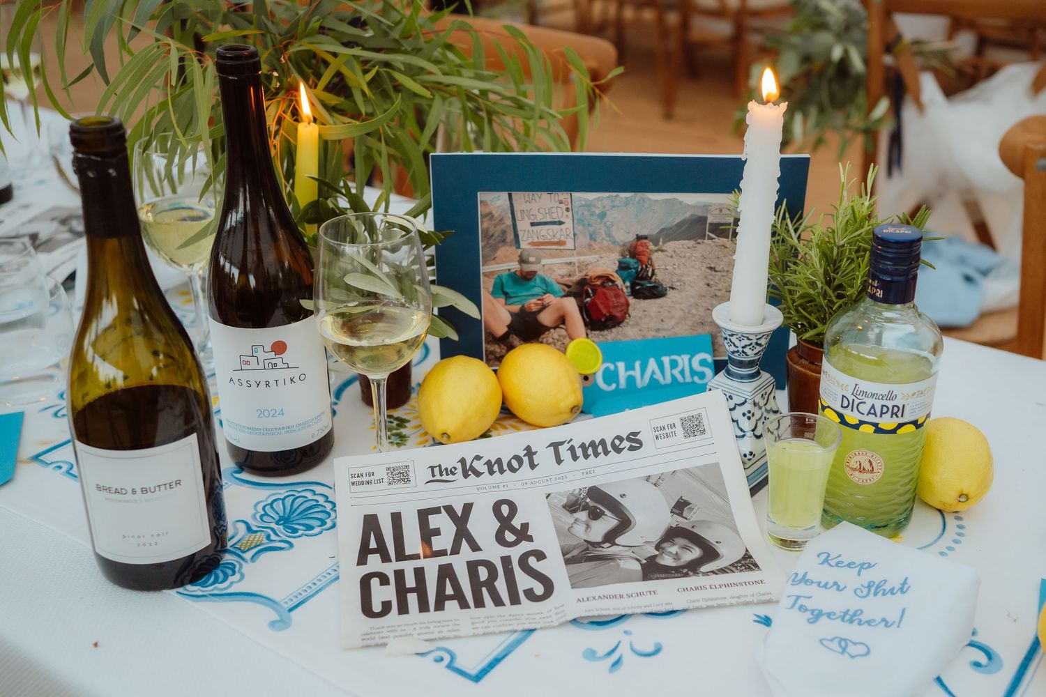 Wedding reception table decorated with wine bottles, newspaper-style signage, and citrus fruits on a blue tablecloth.