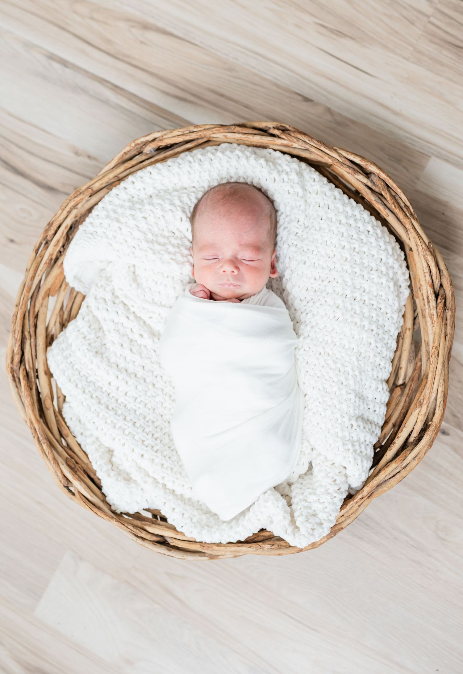 A tiny baby swaddled in white rests on a cream-colored crocheted blanket while a fluffy dog peeks into frame.