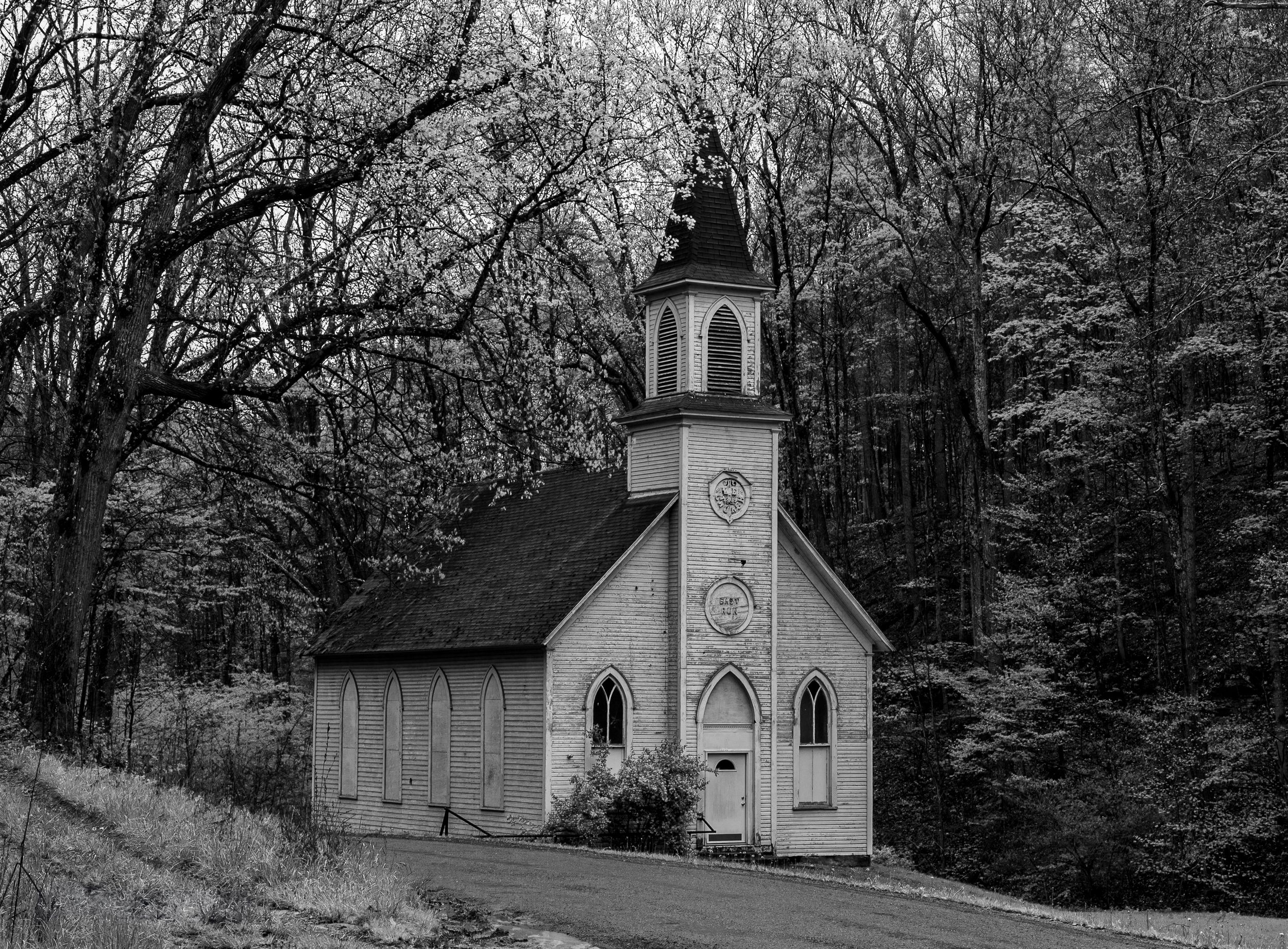 A small white country church with a steeple stands nestled among bare trees in a black and white photograph.