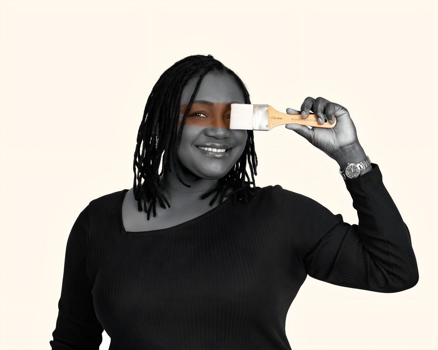 Smiling woman in black outfit holds a paint brush over one eye against a light neutral background.