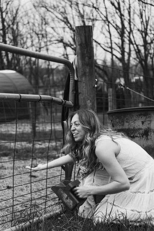 A person in a white dress lies down by a rustic fence in a black and white countryside scene.