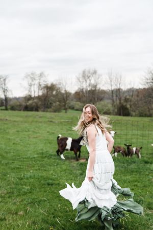 A person in a flowing white dress runs through a green pasture with grazing cows in the background.