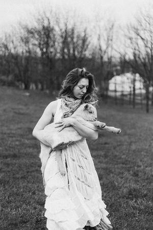 A person in a flowing white dress holds a small lamb while standing in a grassy field on an overcast day.