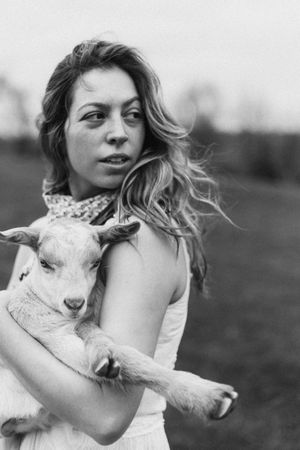 Black and white portraits show a woman in a field holding and cuddling with a small white goat.