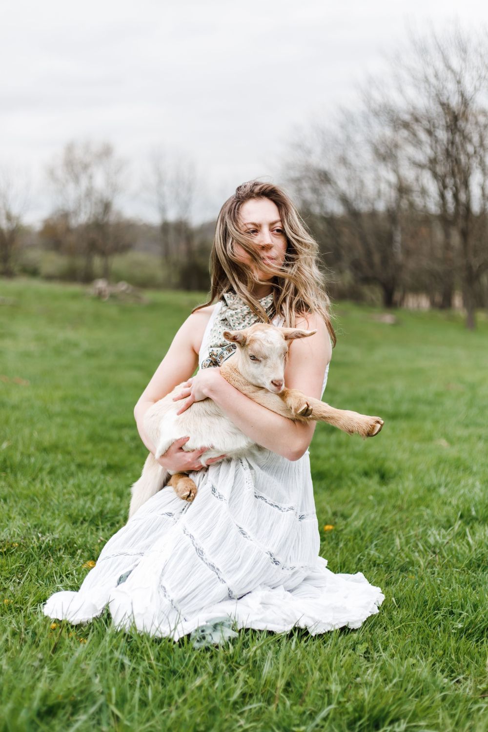 A person in a flowing white dress sits in a green meadow holding a fluffy cat while wind blows through their hair.