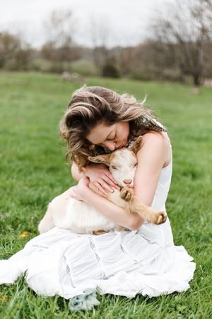 A person in a white dress lies in a green grass field with a dog on a sunny day.