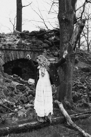 A black and white photo shows a stone wall in the woods with a figure in a white dress standing near trees.