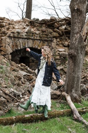 A person in a white dress and denim jacket walks along a rustic stone wall in a wooded outdoor setting.