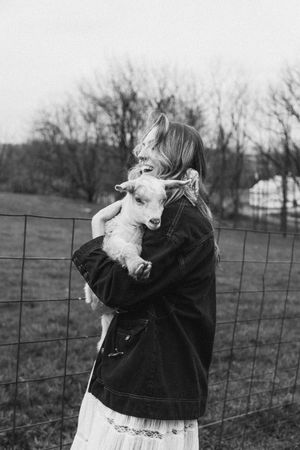 A baby goat wearing a black jacket stands near a wire fence in a rural field setting.