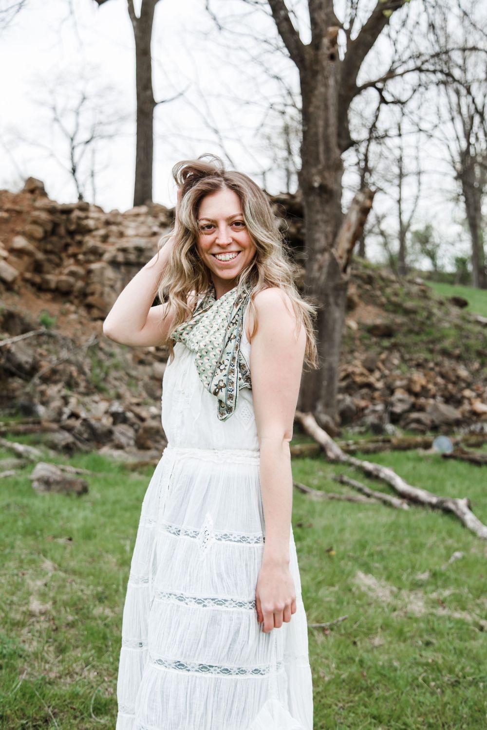 A person wearing a white lace dress poses joyfully in front of bare trees and fallen leaves.