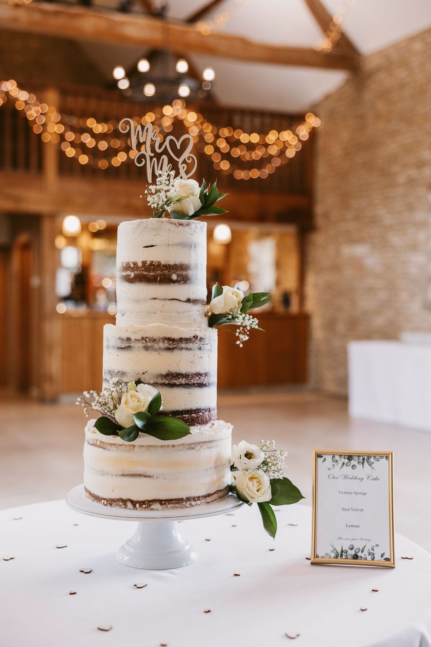 A three-tiered semi-naked wedding cake with white frosting and floral decorations displayed in a rustic venue with string lights.