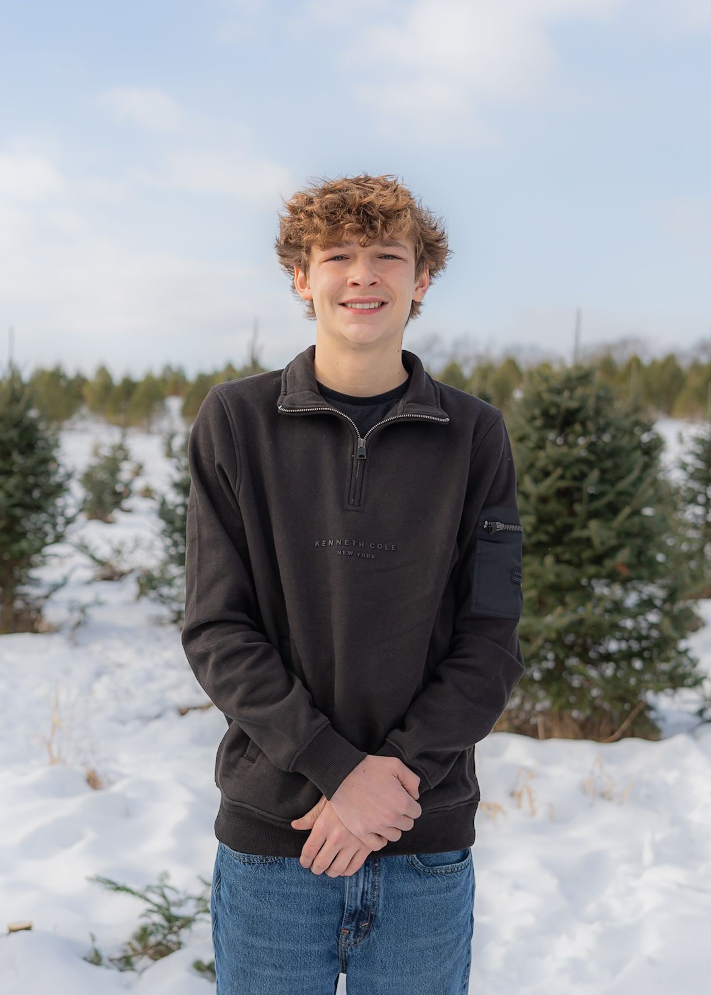 A person in a black sweater and jeans poses in a snowy winter landscape with evergreen trees in the background.