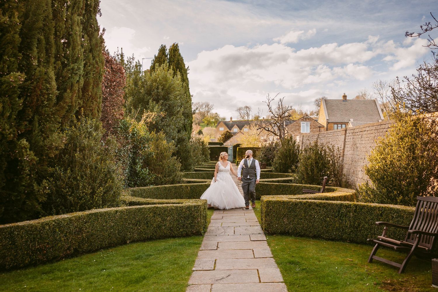 A scenic stone pathway leads through a formal garden with manicured hedges and benches on an autumn day.
