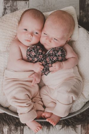 Two infants laying together on a white cushion wearing matching outfits with a floral heart detail.