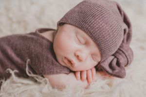 Sleeping baby wearing a purple knit hat and sweater on soft white fabric.