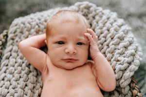 Baby rests on chunky knit blanket while touching head with hands.
