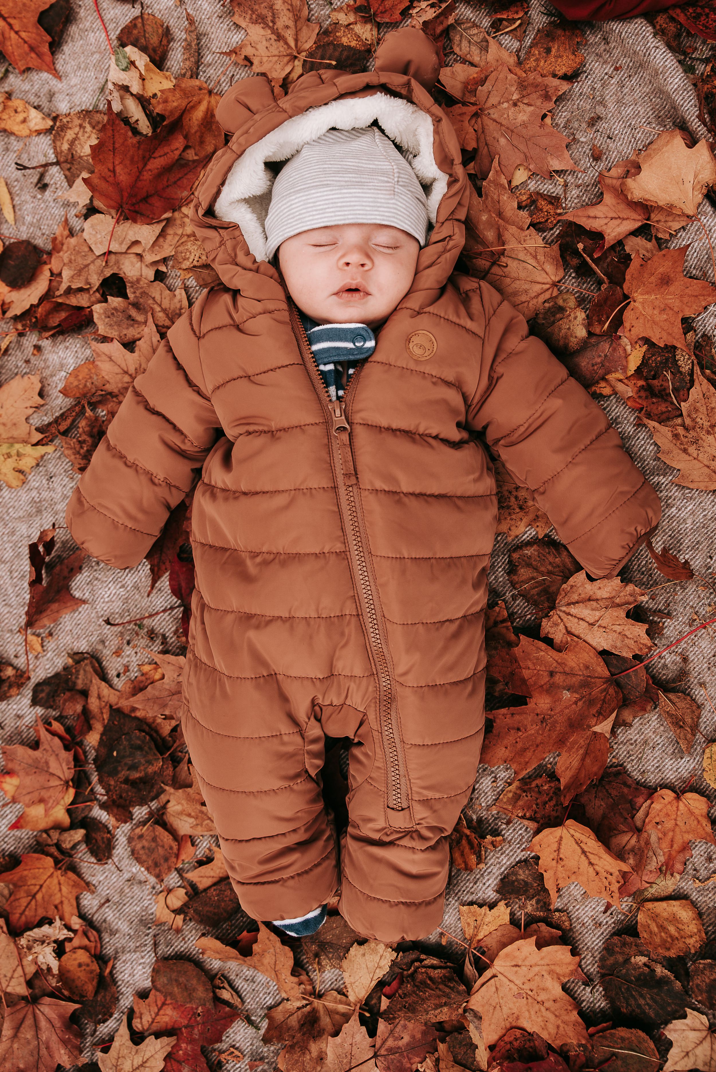 Baby in brown snowsuit sleeping peacefully among autumn leaves.