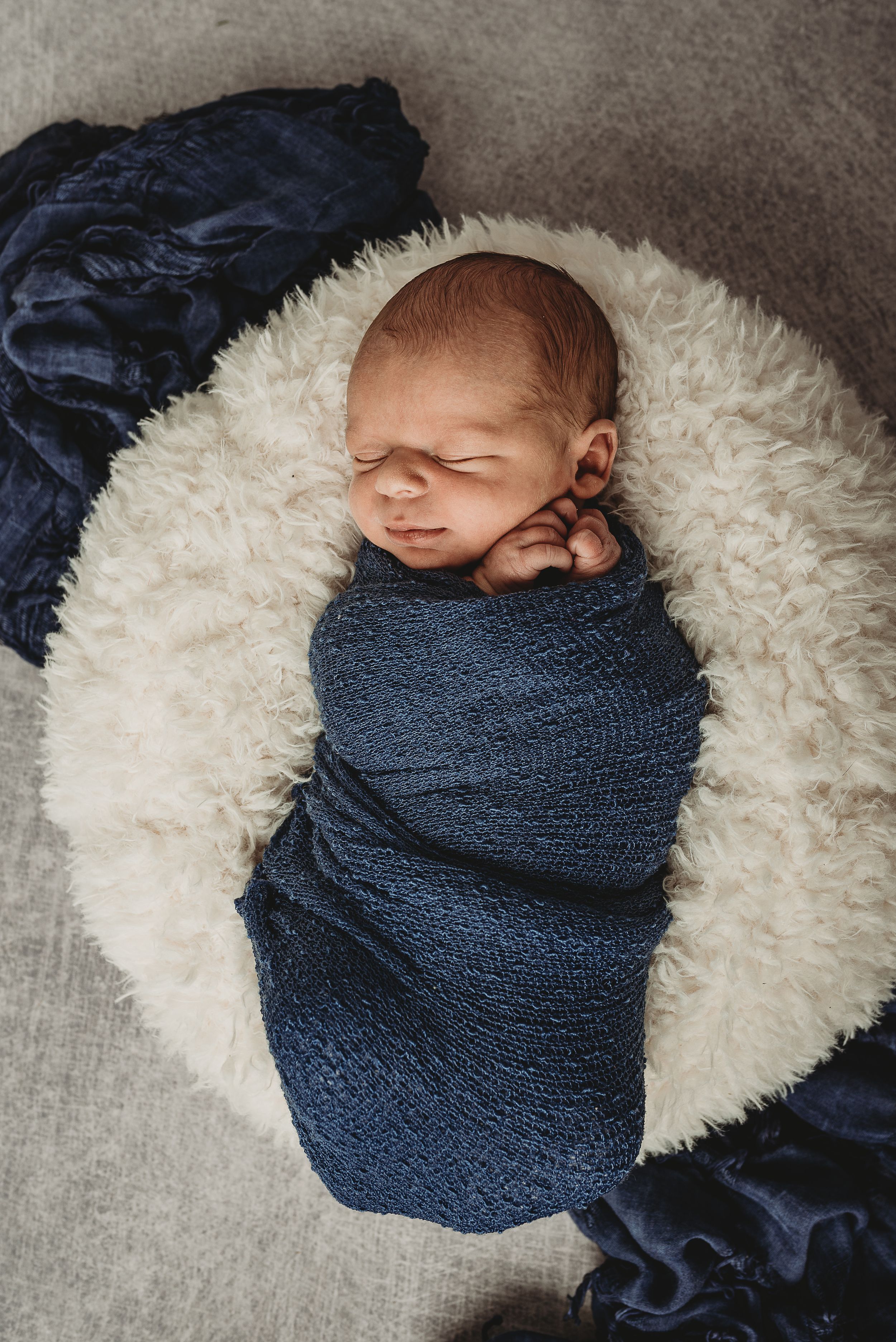 Sleeping newborn wrapped in navy blue swaddle resting on white fluffy blanket.