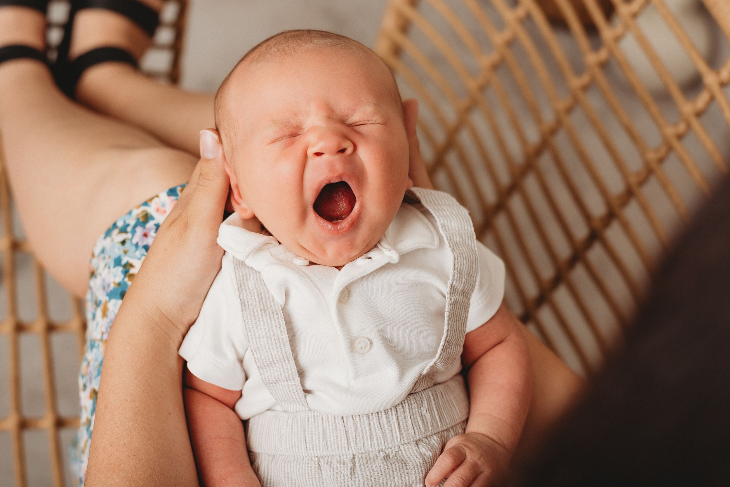 A yawning infant wearing white clothes rests on a wicker chair.