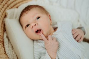 Baby in white knit outfit laying in woven basket with finger near mouth.