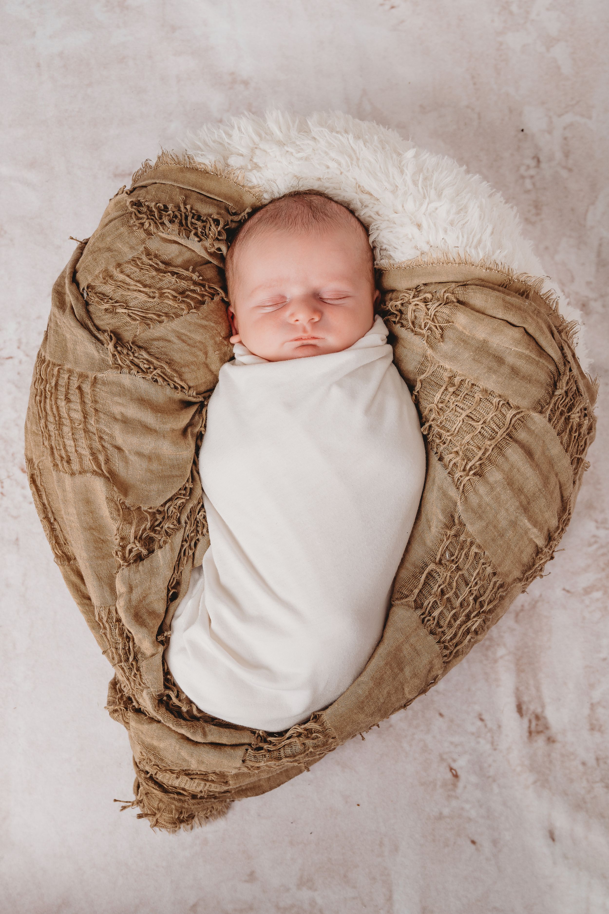 Sleeping newborn wrapped in white swaddle on textured brown heart-shaped backdrop.