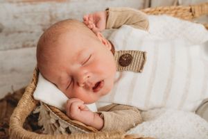 Sleeping newborn in white swaddle rests on tan textured blanket in woven basket.