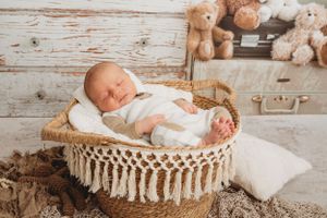Newborn baby sleeping peacefully in macrame basket with teddy bears in background.