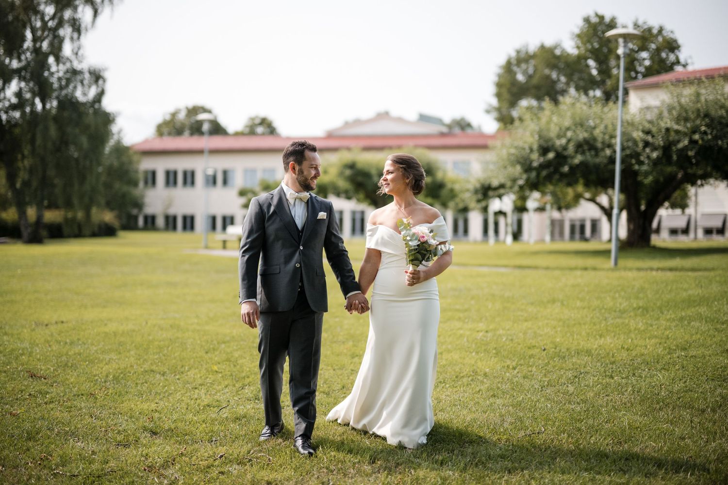 Newlyweds walk hand in hand across a sunlit grassy lawn with white buildings in the background.