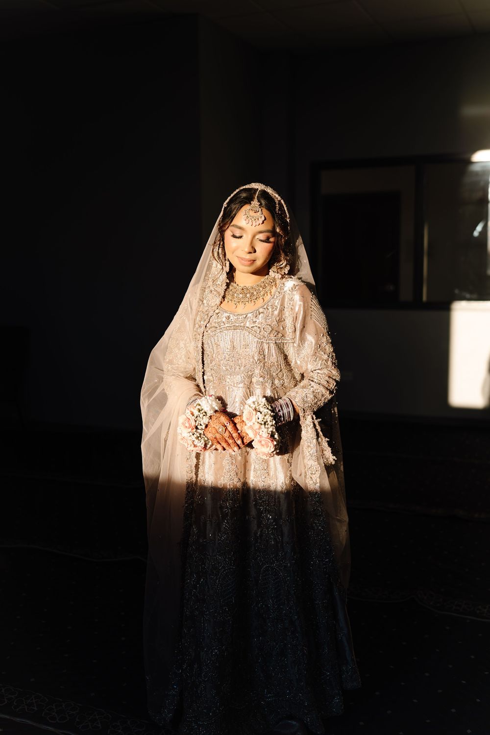 South Asian bride in ivory embroidered lehenga holding flower bouquet in dramatic natural light.