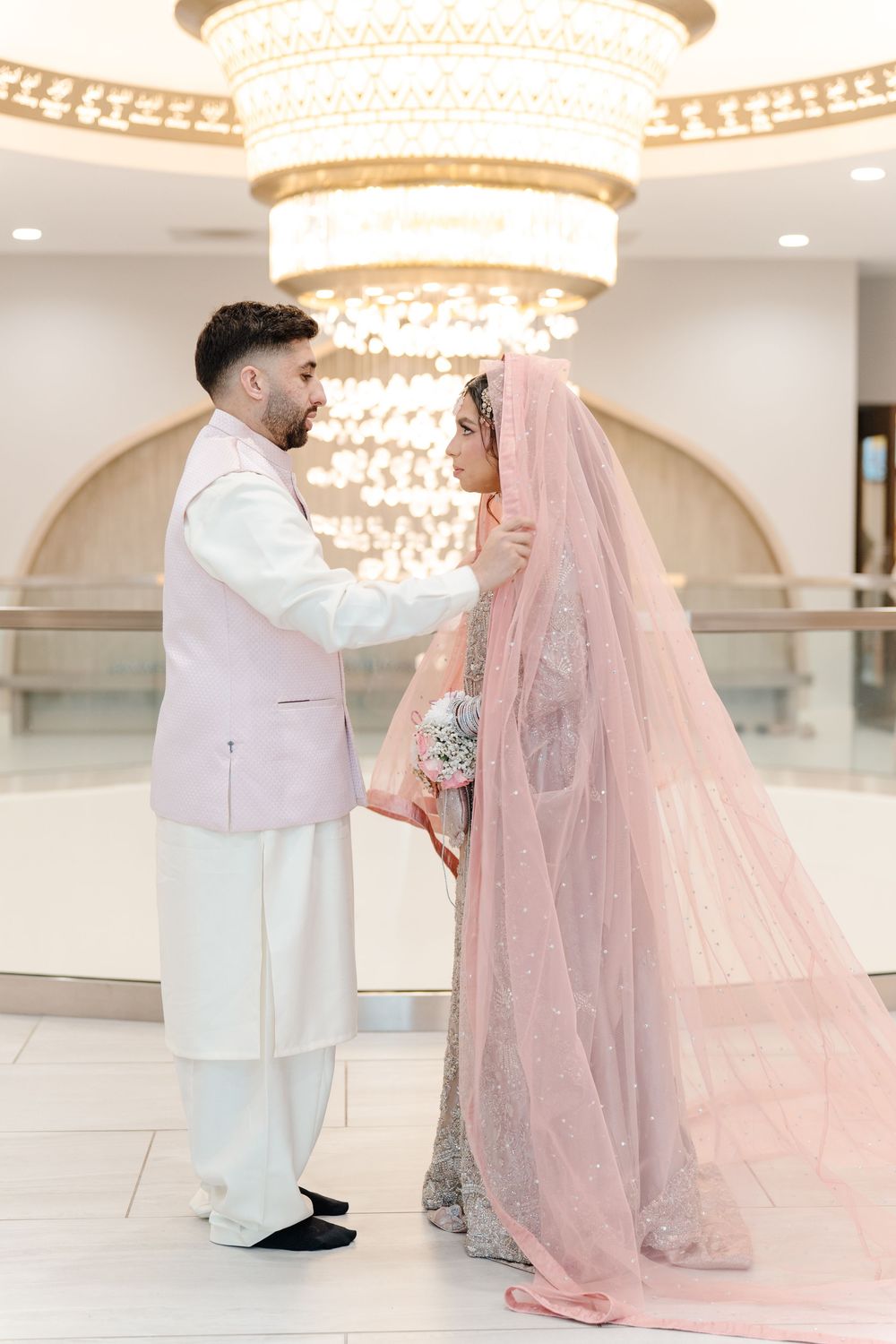 South Asian bride and groom in pink and white traditional wedding attire share a tender moment under a chandelier.