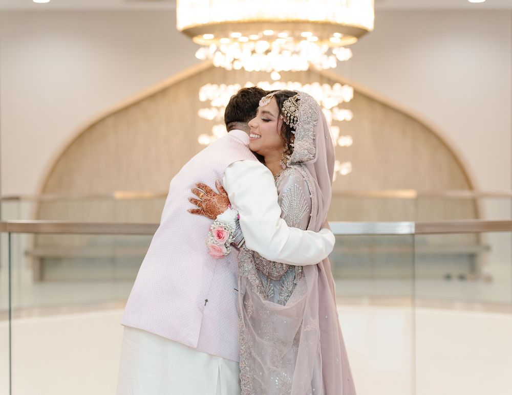 South Asian bride and groom share a joyful embrace under a chandelier in traditional wedding attire.