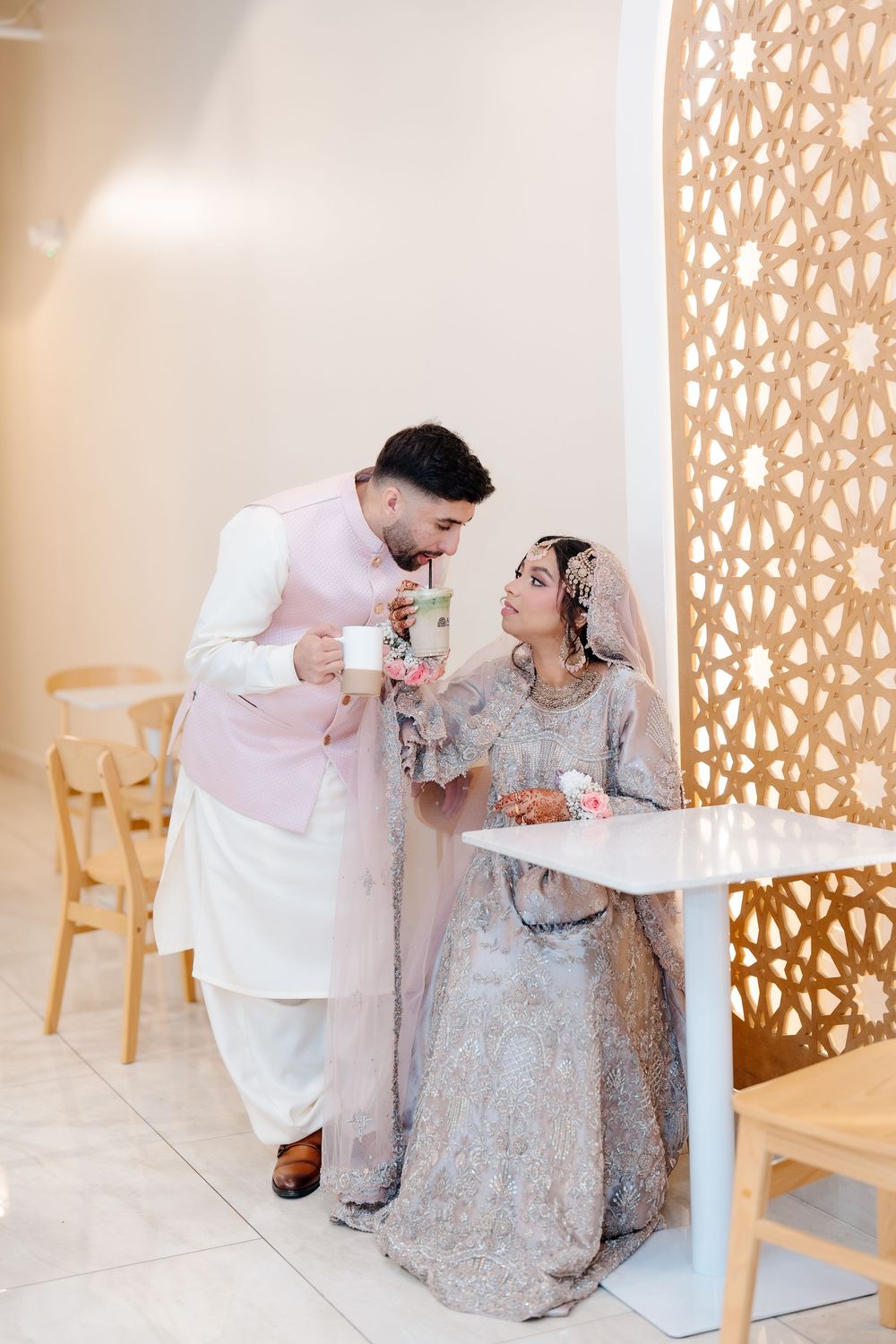 South Asian groom shares a drink with his bride in traditional wedding attire at a cafe setting.