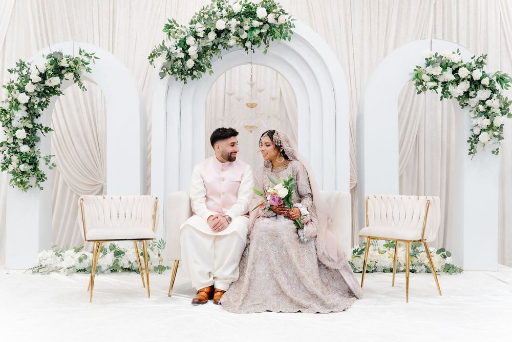 South Asian bride and groom sit together at a white floral wedding stage in elegant traditional attire.