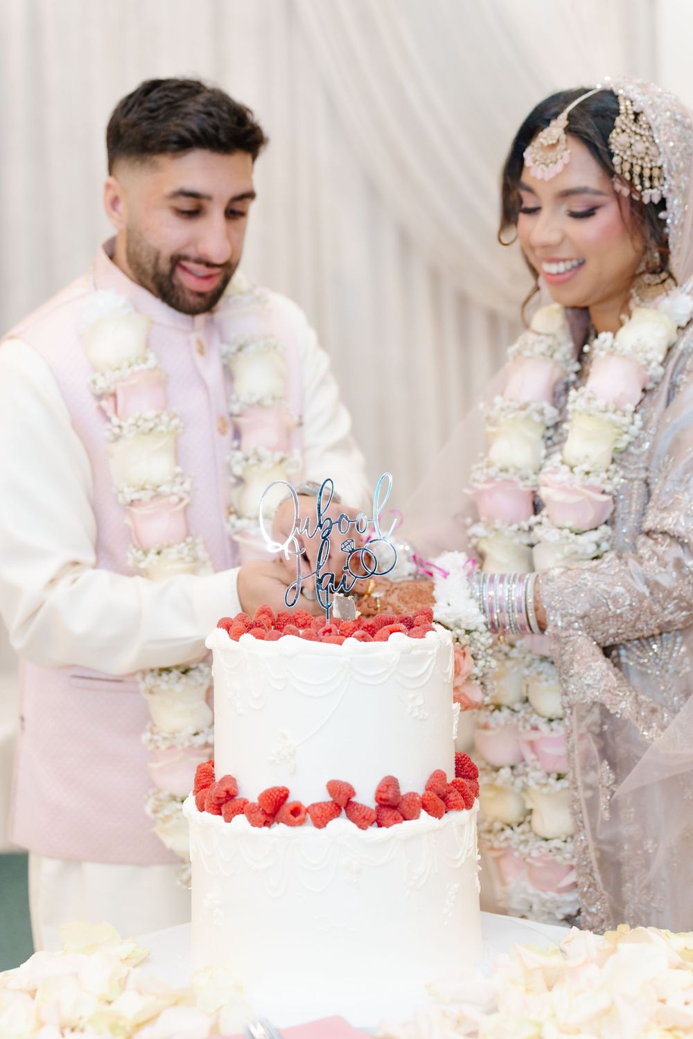 South Asian bride and groom cut a two-tier white wedding cake adorned with fresh raspberries and floral garlands.