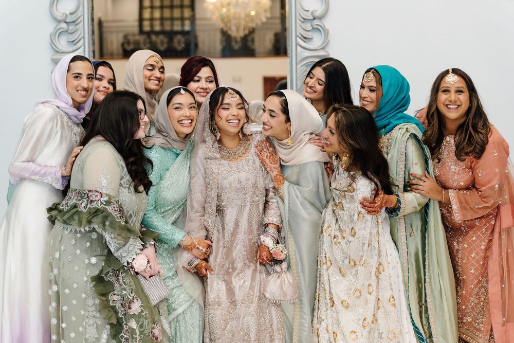 South Asian bride surrounded by smiling bridesmaids in pastel traditional outfits at a wedding celebration.