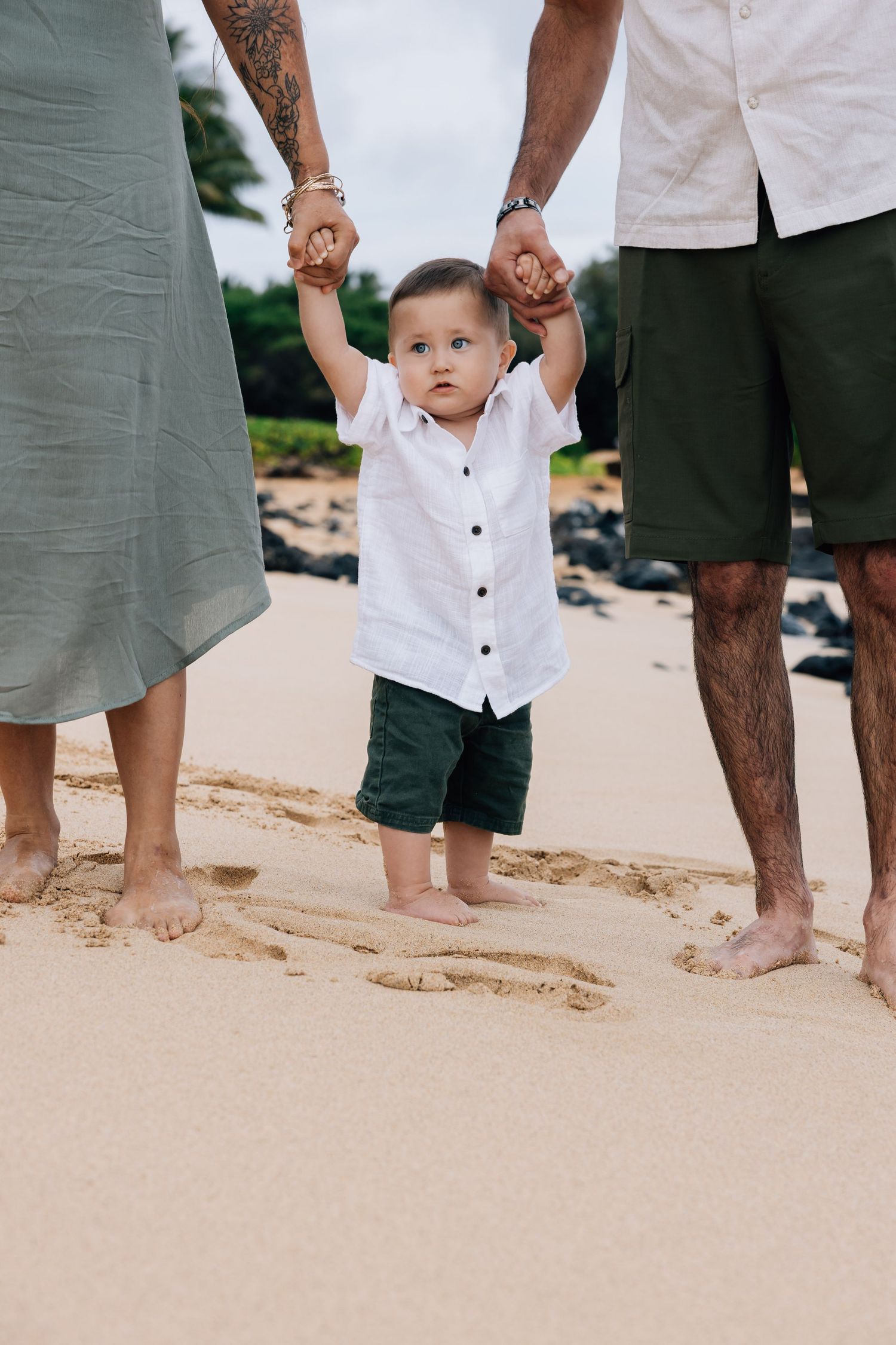 Curious toddler holding parents hands while standing barefoot on sandy beach.