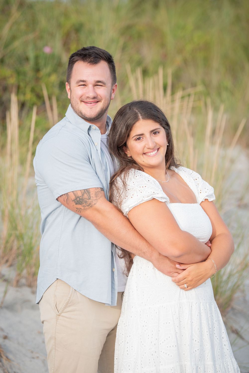 A couple poses for a romantic beach portrait in casual attire against tall grass.