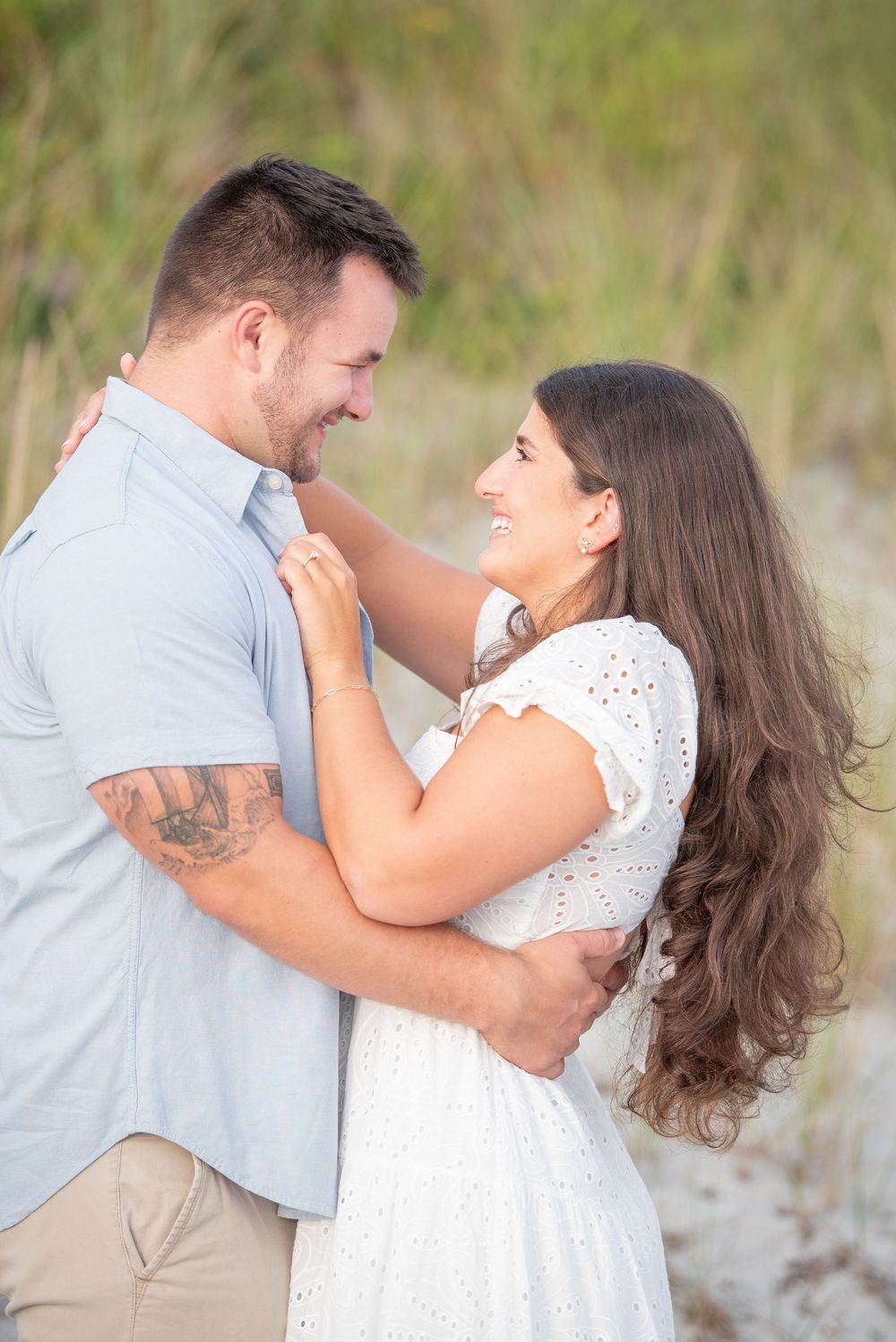 A couple shares a tender moment on the beach while wearing coordinating light-colored outfits.
