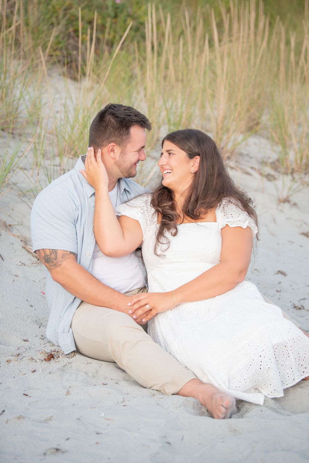 A couple sits together on sandy beach dunes during a romantic photoshoot.