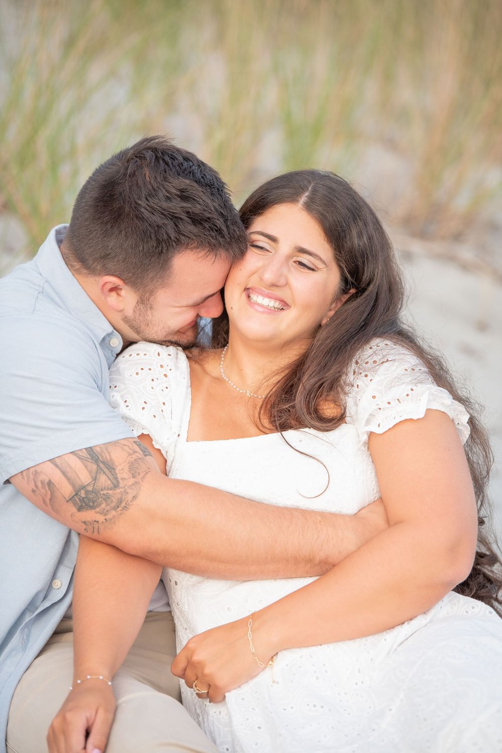 A joyful couple embraces and laughs together on a sandy beach with tall grass in the background.