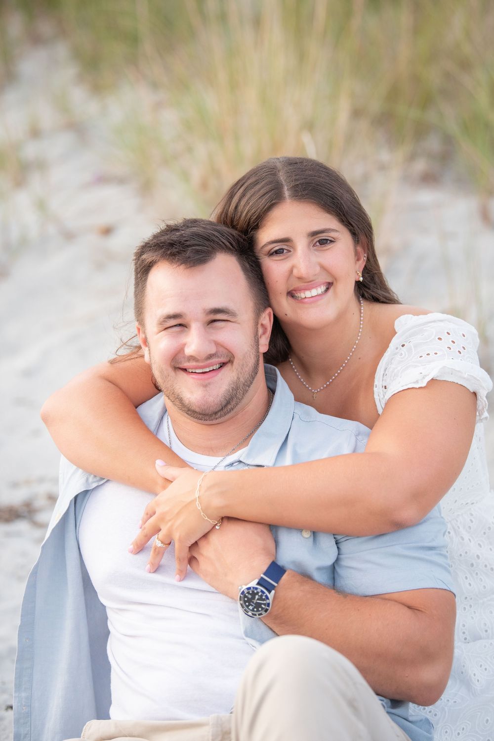 A happy couple embraces while sitting on beach dunes during a photo session.