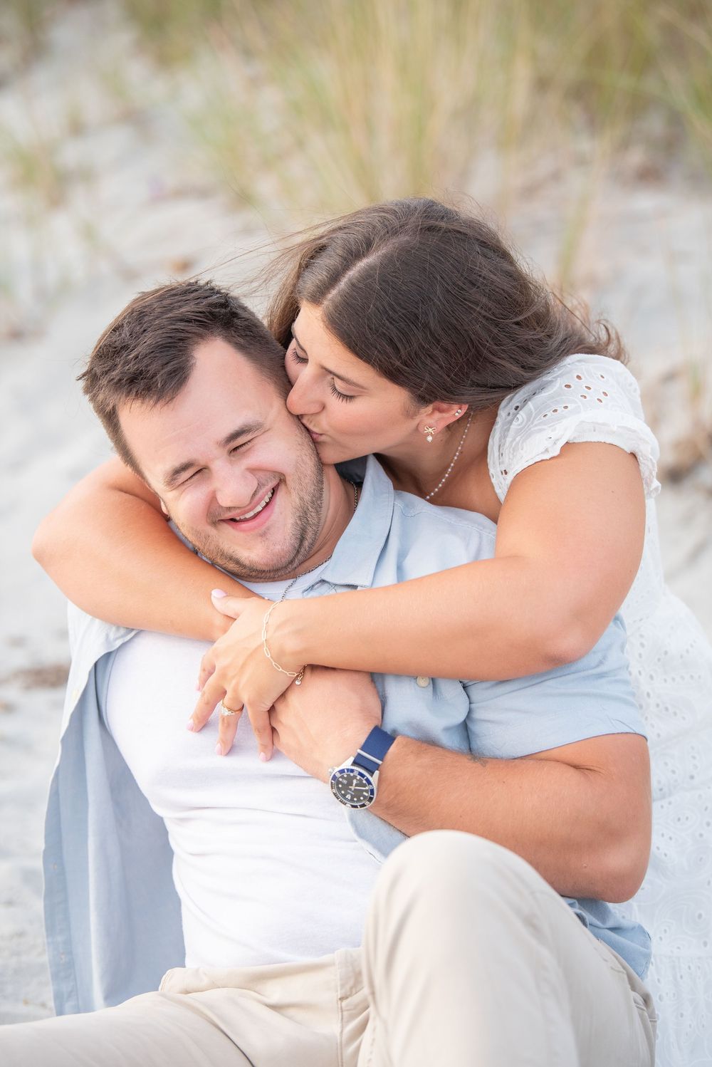 A couple shares a playful moment of affection on the beach with sea grass in the background.