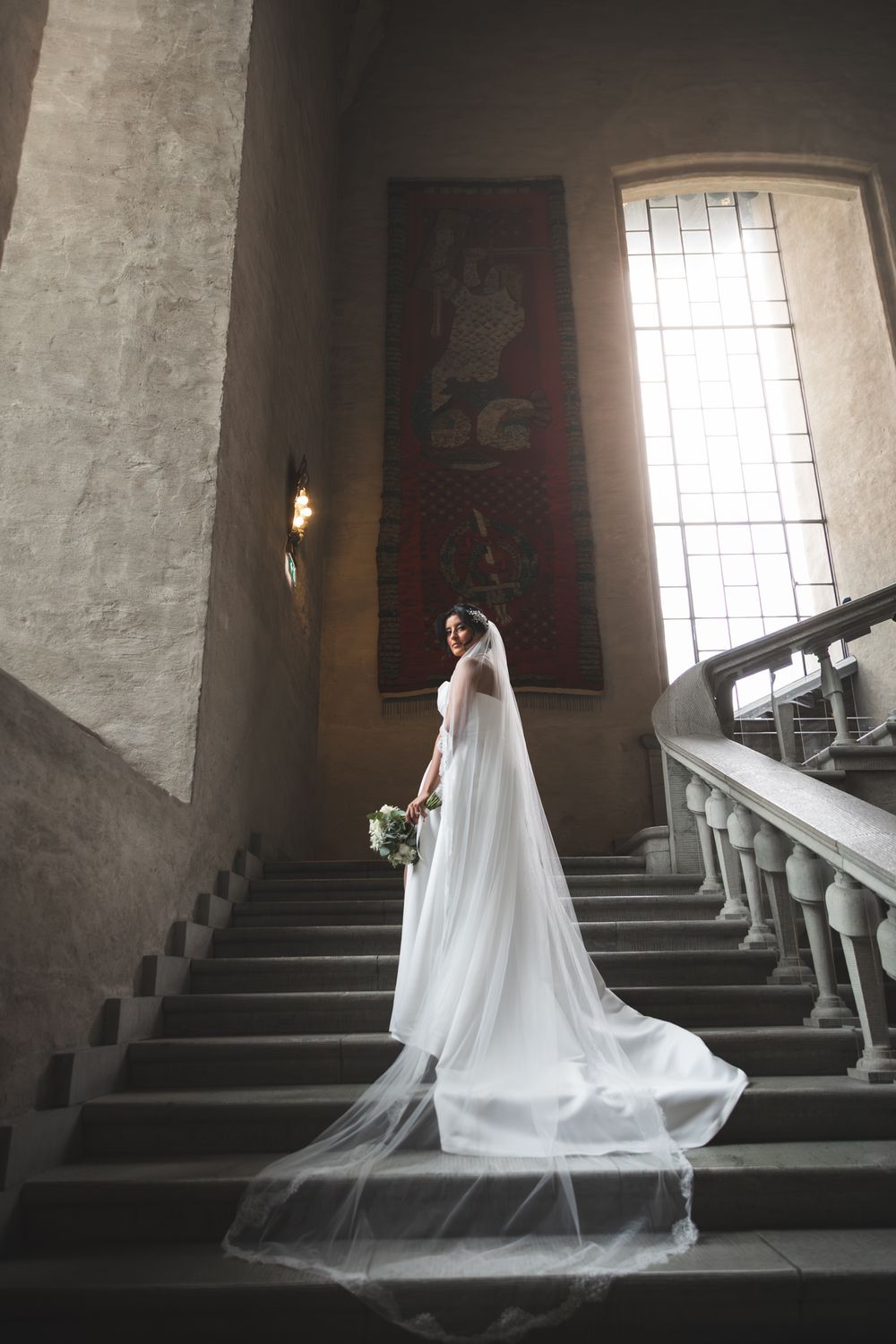 A flowing white wedding gown and veil creates an elegant silhouette against stone stairs in dramatic lighting.