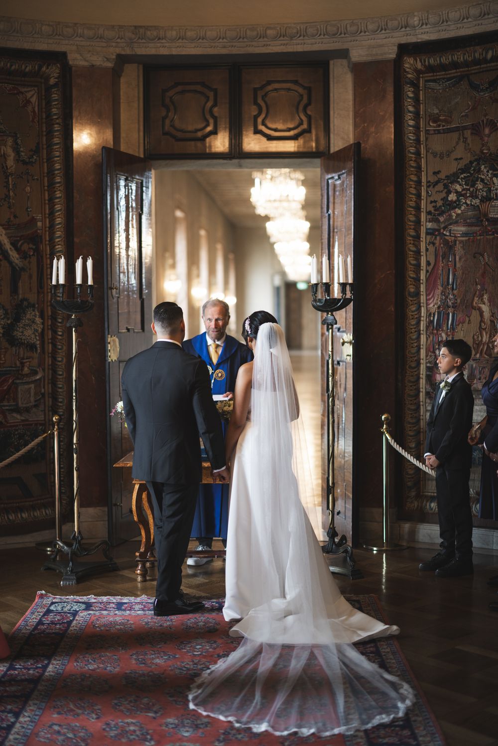 Wedding ceremony taking place in an elegant room with ornate doors, chandelier lighting and a long flowing veil.