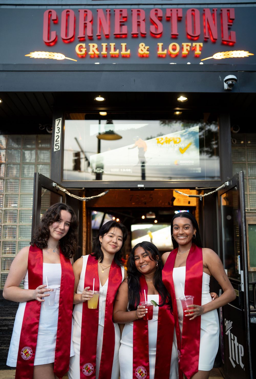 Graduates in matching red and white attire posing at Cornerstone Grill entrance.