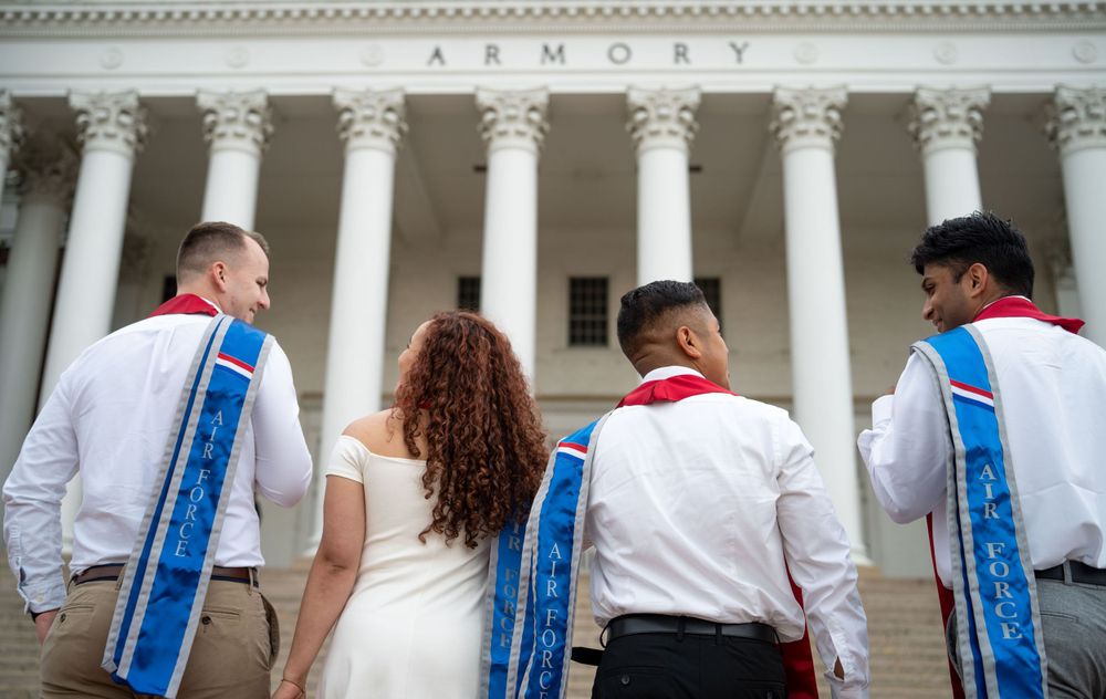 Row of graduates in formal attire standing in front of classical columned building.