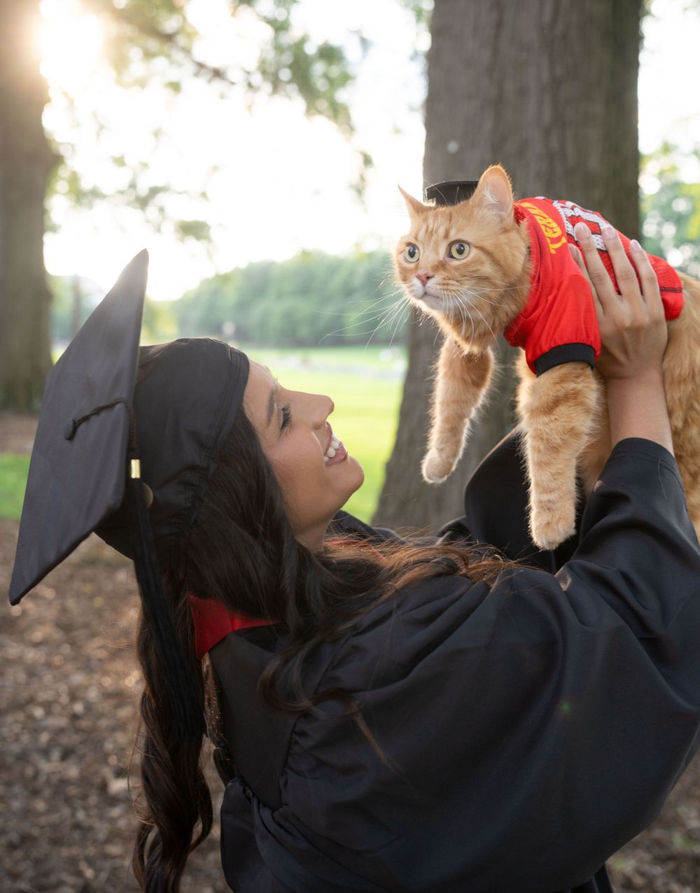 Graduate in academic regalia holding orange cat while lying on ground outdoors.